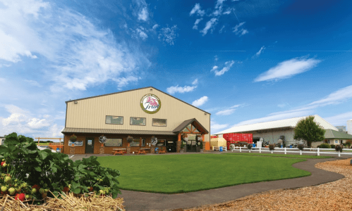 A large farm building with a sign, surrounded by green grass and blue skies, featuring a pathway and nearby structures.