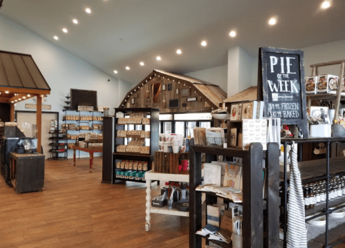 Interior of a cozy shop featuring shelves of products, a display for "Pie of the Week," and a rustic house structure.