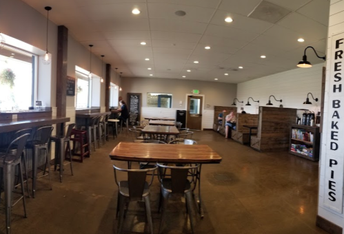 Interior of a cozy café with wooden tables, metal chairs, and a sign for "Fresh Baked Pies." Natural light from windows.