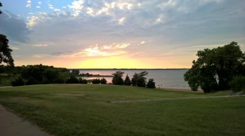 A serene lakeside view at sunset, with grassy areas and trees under a partly cloudy sky.