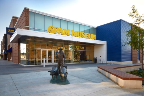 Exterior of the Spam Museum featuring a large sign and a bronze statue of a man with two pigs in front.
