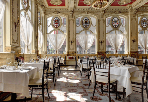 Elegant restaurant interior with large windows, ornate decor, and neatly set tables. Bright and inviting atmosphere.