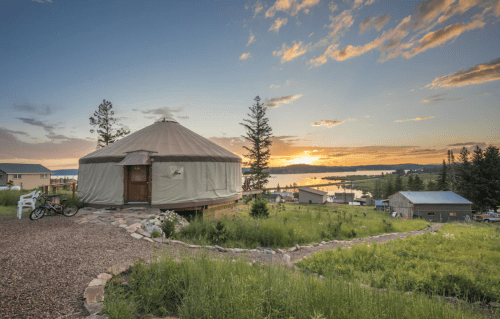 A yurt on a hillside at sunset, surrounded by trees and overlooking a lake with distant buildings.