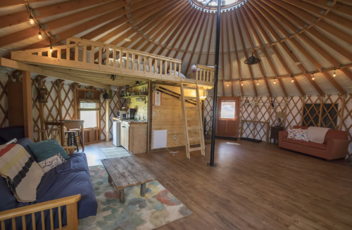 Interior of a cozy yurt with a loft, wooden furniture, and warm lighting, featuring a living area and kitchenette.