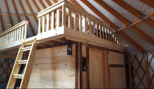 Interior of a wooden yurt featuring a loft area with a railing and a ladder, showcasing rustic design elements.