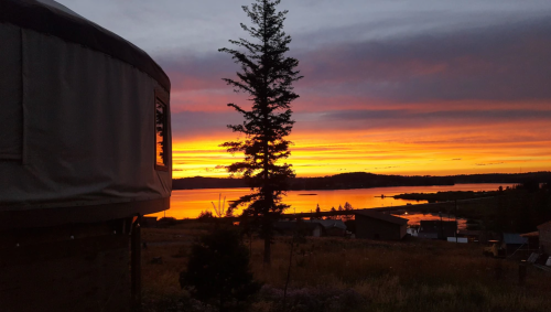 A serene sunset over a lake, with a silhouette of a tree and a yurt in the foreground. Vibrant orange and purple skies.