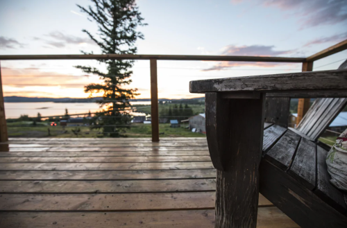 A wooden deck with a chair overlooks a scenic sunset by a lake, framed by trees and distant buildings.