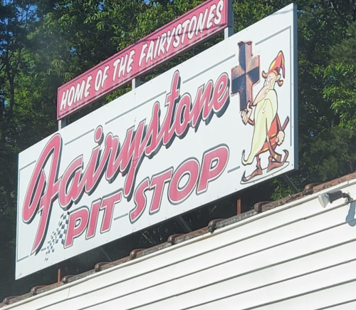 Sign for "Fairystone Pit Stop" featuring a cartoon character holding a cross, surrounded by trees.
