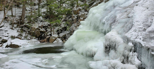A frozen waterfall surrounded by snow-covered rocks and trees, creating a serene winter landscape.