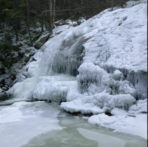 A frozen waterfall surrounded by snow-covered rocks and icy terrain in a winter forest setting.
