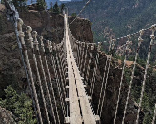 A long suspension bridge stretches over a rocky canyon, surrounded by trees and mountains in the background.