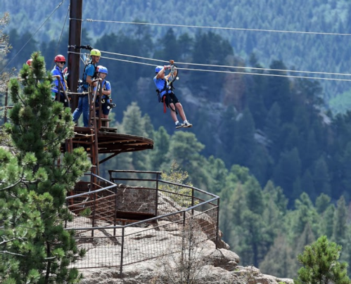 A person zip-lining from a platform, with others watching from a nearby lookout surrounded by trees and mountains.