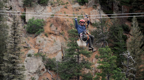 A person in safety gear zip-lining across a canyon, surrounded by trees and rocky terrain.