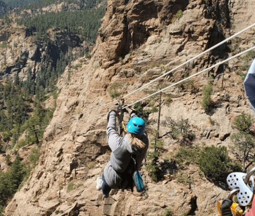 A person zip-lining over a rocky landscape with trees in the background, wearing a helmet and harness.
