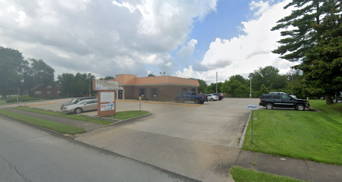 A brick building with a metal roof, surrounded by a parking lot and green grass under a partly cloudy sky.