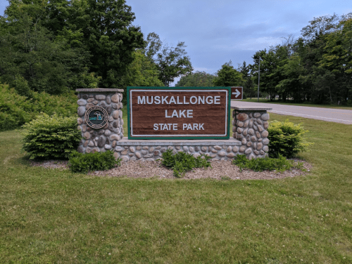 Sign for Muskallonge Lake State Park, featuring stone base and greenery, with a road visible in the background.