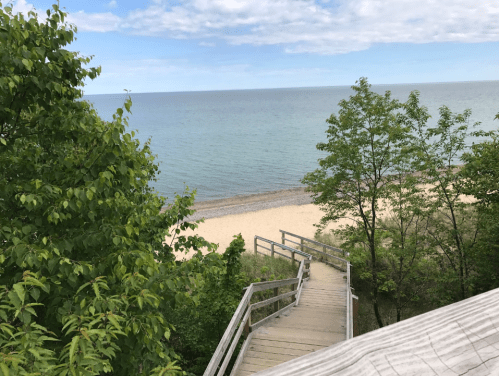 A wooden staircase leads down to a sandy beach by a calm lake, surrounded by green trees and a clear blue sky.
