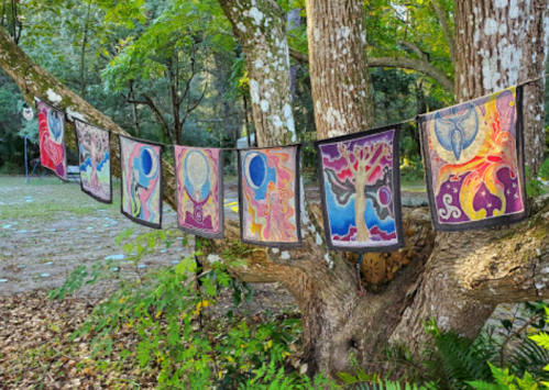 Colorful fabric banners with intricate designs hang from a tree in a lush, green outdoor setting.