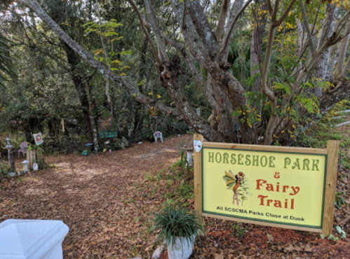 Sign for Horseshoe Park & Fairy Trail, surrounded by trees and fallen leaves, inviting visitors to explore.