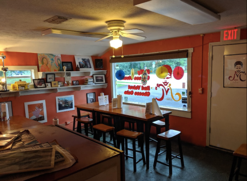 Cozy interior of a cafe with orange walls, wooden tables, and artwork displayed on shelves. Brightly lit with a ceiling fan.