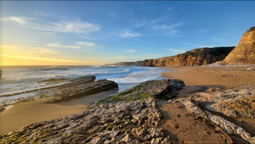 A serene beach at sunset, with rocky formations, gentle waves, and cliffs in the background under a clear blue sky.