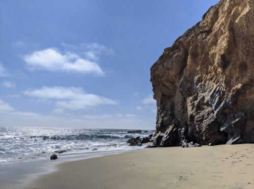 A sandy beach with gentle waves, rocky cliffs, and a clear blue sky with scattered clouds.