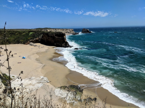 A scenic beach with golden sand, gentle waves, and cliffs under a clear blue sky. People enjoy the sun and water.