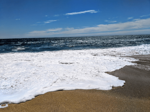 Waves gently wash onto a sandy beach under a clear blue sky, creating white foam along the shoreline.