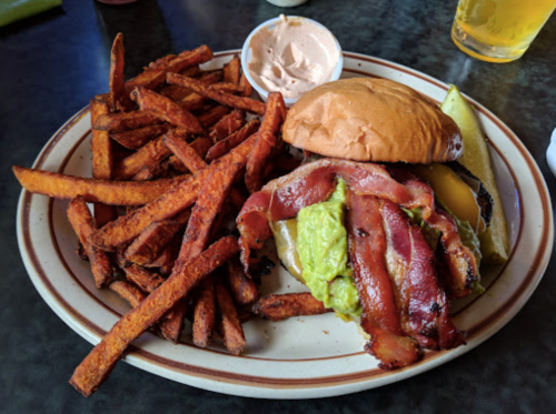 A juicy burger topped with bacon and avocado, served with crispy sweet potato fries and a dipping sauce.