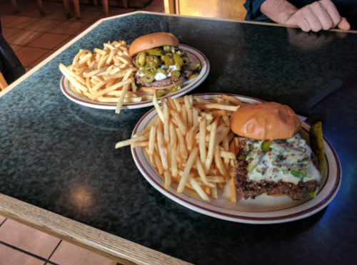 Two plates on a table: one with a burger topped with pickles and onions, the other with a cheeseburger, both served with fries.