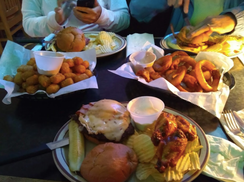A table with various dishes: burgers, onion rings, chicken, and tater tots, with sides and dipping sauces.