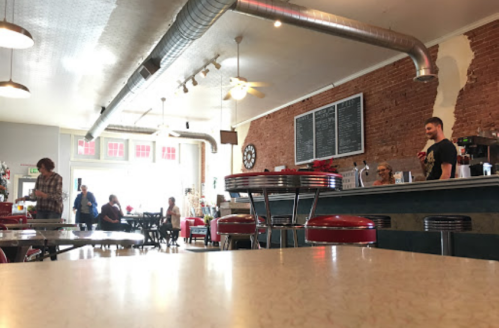 A cozy diner with red stools, brick walls, and patrons enjoying their meals. A staff member serves at the counter.