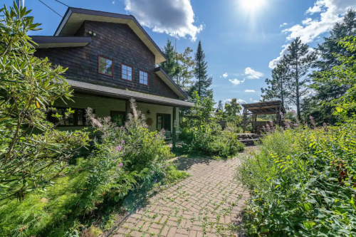 A charming house surrounded by lush greenery and colorful flowers, with a sunny sky and a patio area in the background.