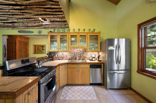 A cozy kitchen with wooden cabinets, stainless steel appliances, and a stone backsplash, featuring natural light and rustic decor.