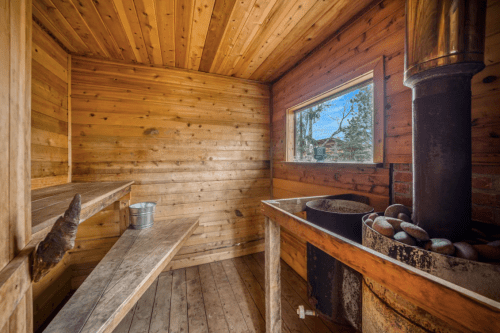 Interior of a wooden sauna with benches, a stove, and a window showing a view of nature outside.