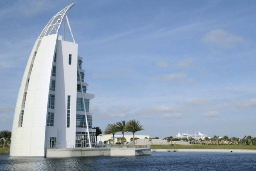 Modern white building with a sail-like design beside a calm water body, surrounded by palm trees and blue sky.