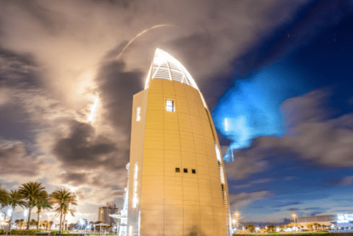 A modern building illuminated at night, surrounded by palm trees and dramatic clouds in the sky.