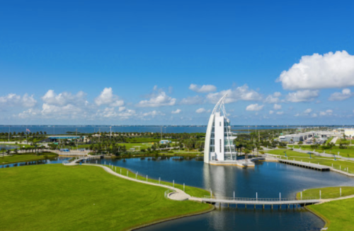 A modern white tower stands by a serene lake, surrounded by green lawns and blue skies with fluffy clouds.
