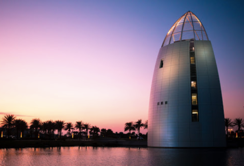 A modern tower with a pointed roof beside a calm body of water, set against a vibrant sunset sky.