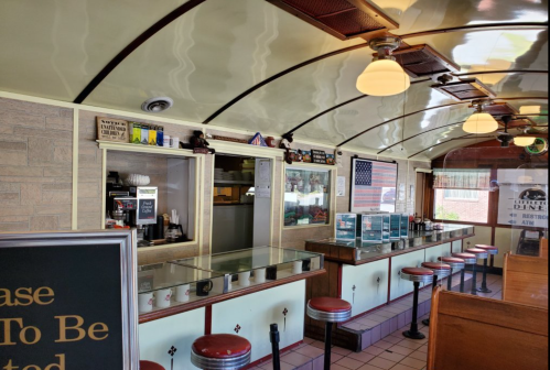 Interior of a retro diner with red stools, a counter, and vintage decor, featuring an American flag in the background.