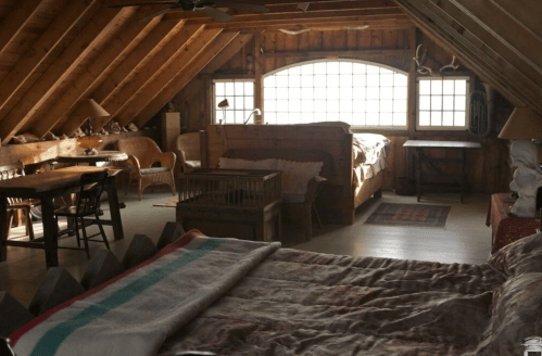 Cozy attic bedroom with wooden beams, two beds, a table, and a warm, rustic atmosphere. Natural light from large windows.