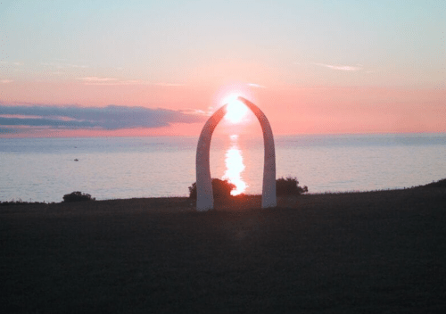 A white arch sculpture silhouetted against a sunset over the ocean, with vibrant colors reflecting on the water.