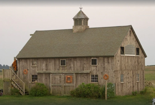 A rustic wooden barn with a cupola, surrounded by greenery and a clear sky in the background.