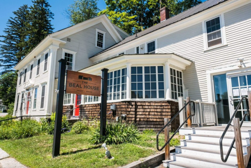 A large, white house with a wooden sign reading "The Beal House" and a welcoming entrance surrounded by greenery.
