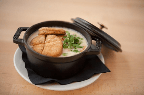 A small black pot filled with creamy soup, topped with herbs and two golden-brown biscuits, on a white plate.
