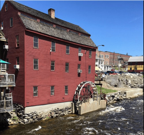 A red mill building beside a flowing river, featuring a water wheel and nearby stone embankment.