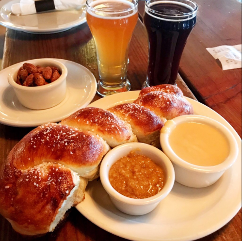 A plate with braided bread, two dipping sauces, a bowl of nuts, and two glasses of beer on a wooden table.