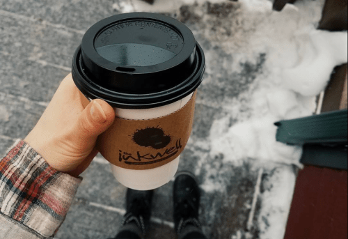 A hand holding a coffee cup with a brown sleeve, snow on the ground, and a plaid shirt visible.
