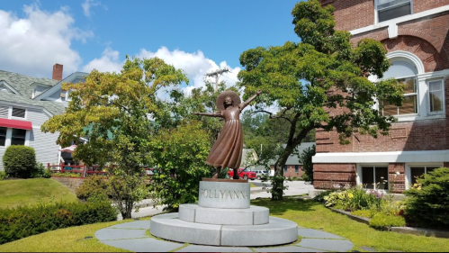 A bronze statue of a girl with outstretched arms, titled "Pollyanna," surrounded by greenery and a brick building.