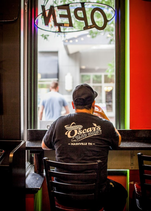 A person sits at a window counter in a restaurant, wearing a black shirt with "Oscar's" printed on it.